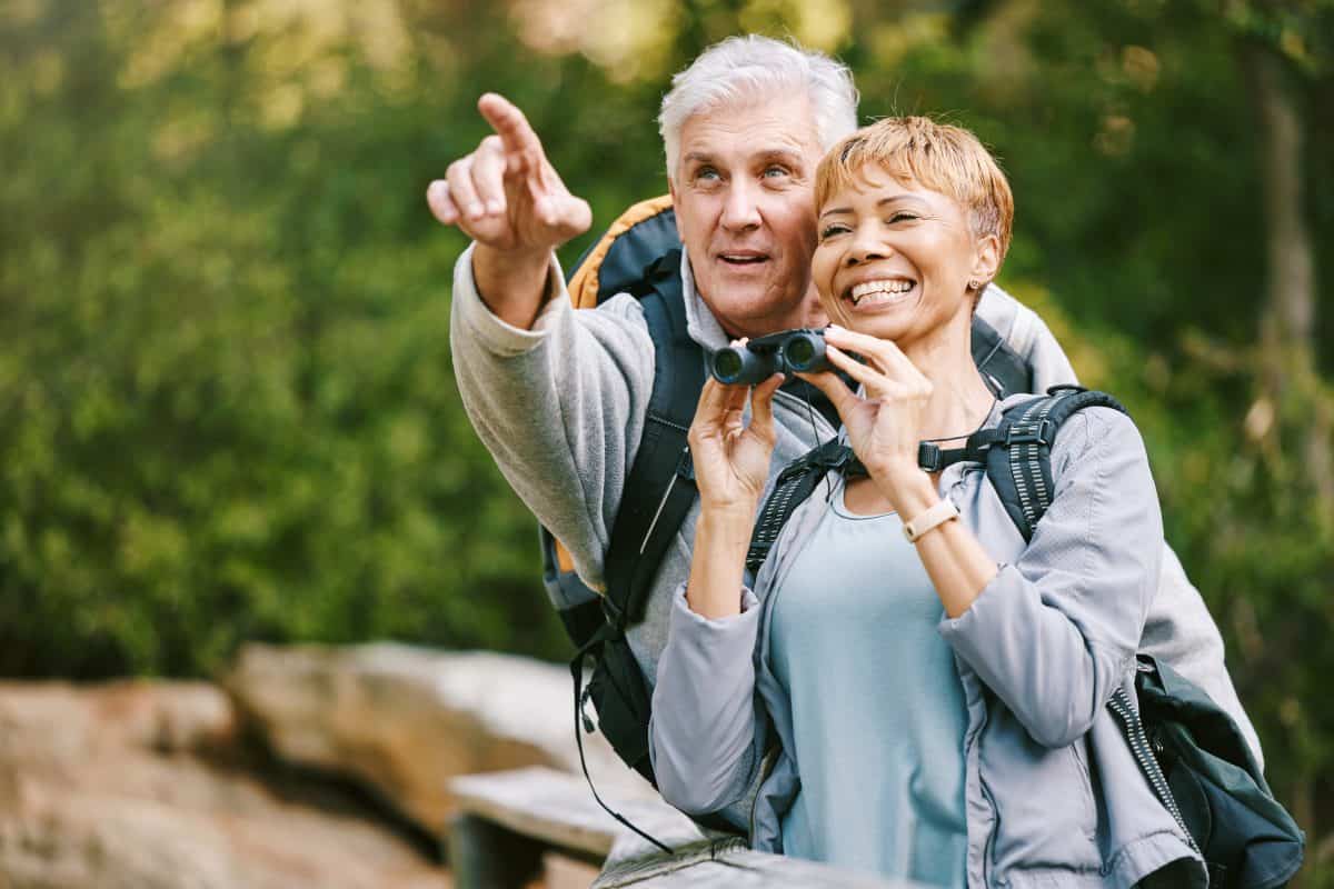 Elderly, couple hiking and bird watching with adventure outdoor, hike together and fitness for active lifestyle. Nature, trekking and senior man pointing and woman with binocular, freedom and travel.