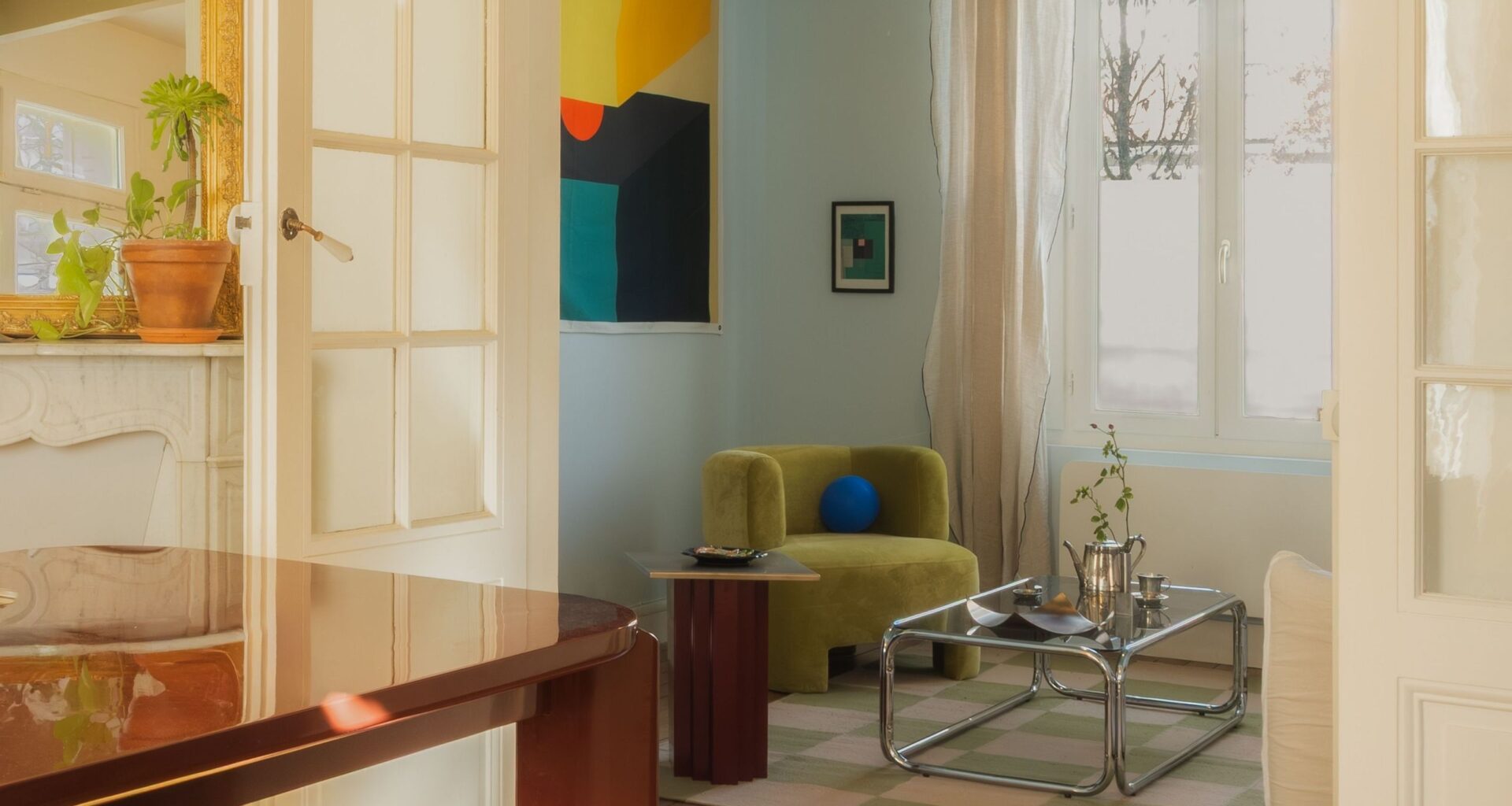 A living room with a lime-toned armchair, abstract colorful artwork, pale blue walls, a tubular chrome and glass coffee table, and white-washed wooden detailing.
