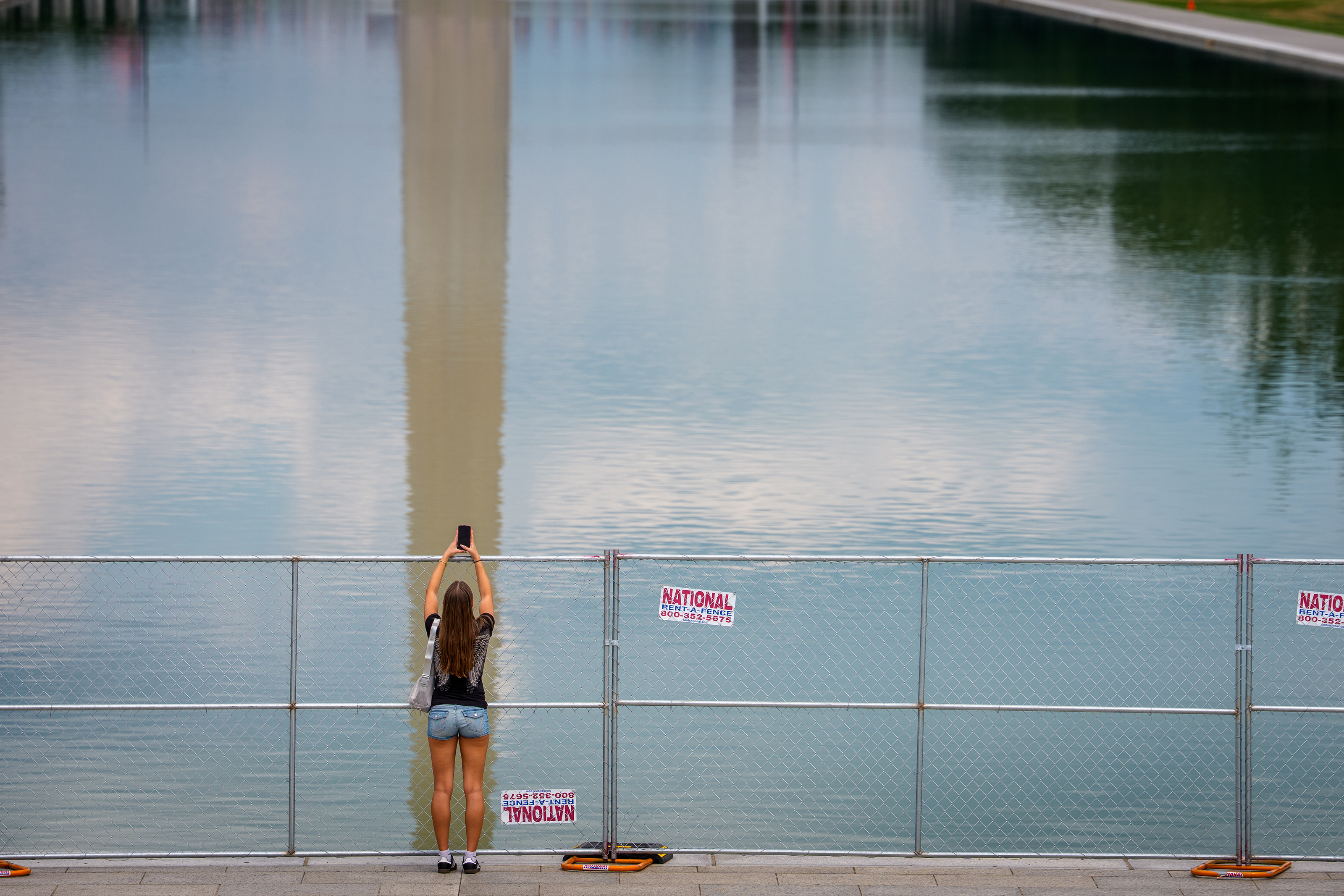 WASHINGTON, DC - JULY 9: The Reflection of the Washington Monument is visible as a visitor takes a picture of the Reflecting Pool though a chain link fence on the National Mall on July 9, 2026 in Washington, DC. Green water remained in the chain link fence enclosed Reflecting Pool on Thursday after Interior Secretary Doug Burgum announced Sunday that crews would drain the pool for a second time in three months for repairs and cleanup following months of maintenance issues, as protesters gathered outside the arraignment hearing of former Olympian David Hearn, who is accused of damaging the landmark. (Photo by Andrew Harnik/Getty Images)