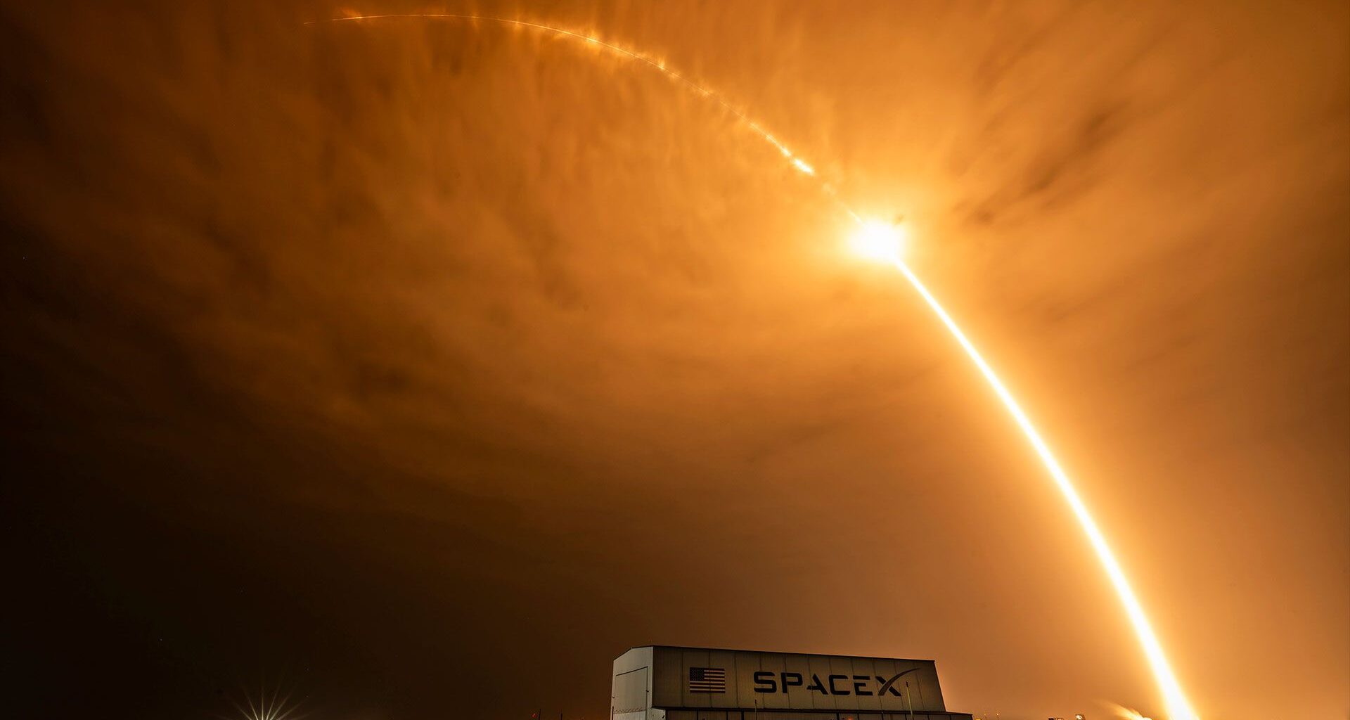 time lapse photo of an early morning rocket launch producing a bright streak arcing over a hangar and stretching beyond the clouds
