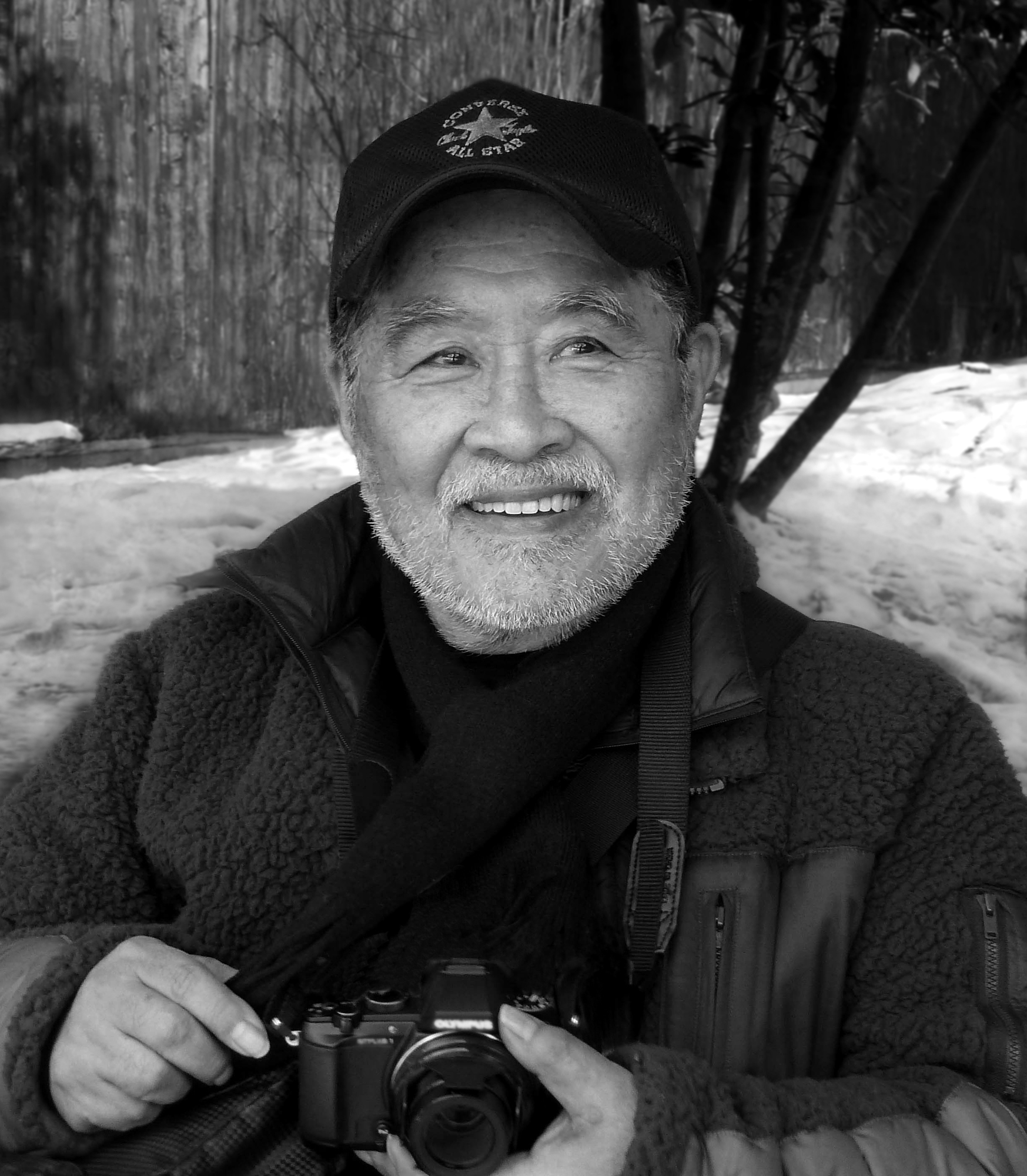 Black and white photo of a smiling elderly man wearing a cap and jacket, holding a camera outdoors in a snowy setting