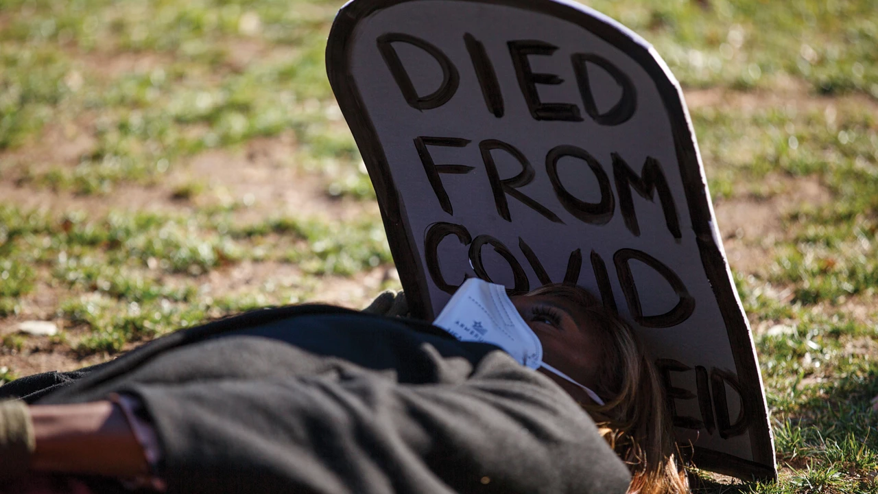 A person wearing a white KN95 mask lies on grassy ground. At their head is a gravestone made from cardboard with the words “DIED FROM COVID” handwritten on it.