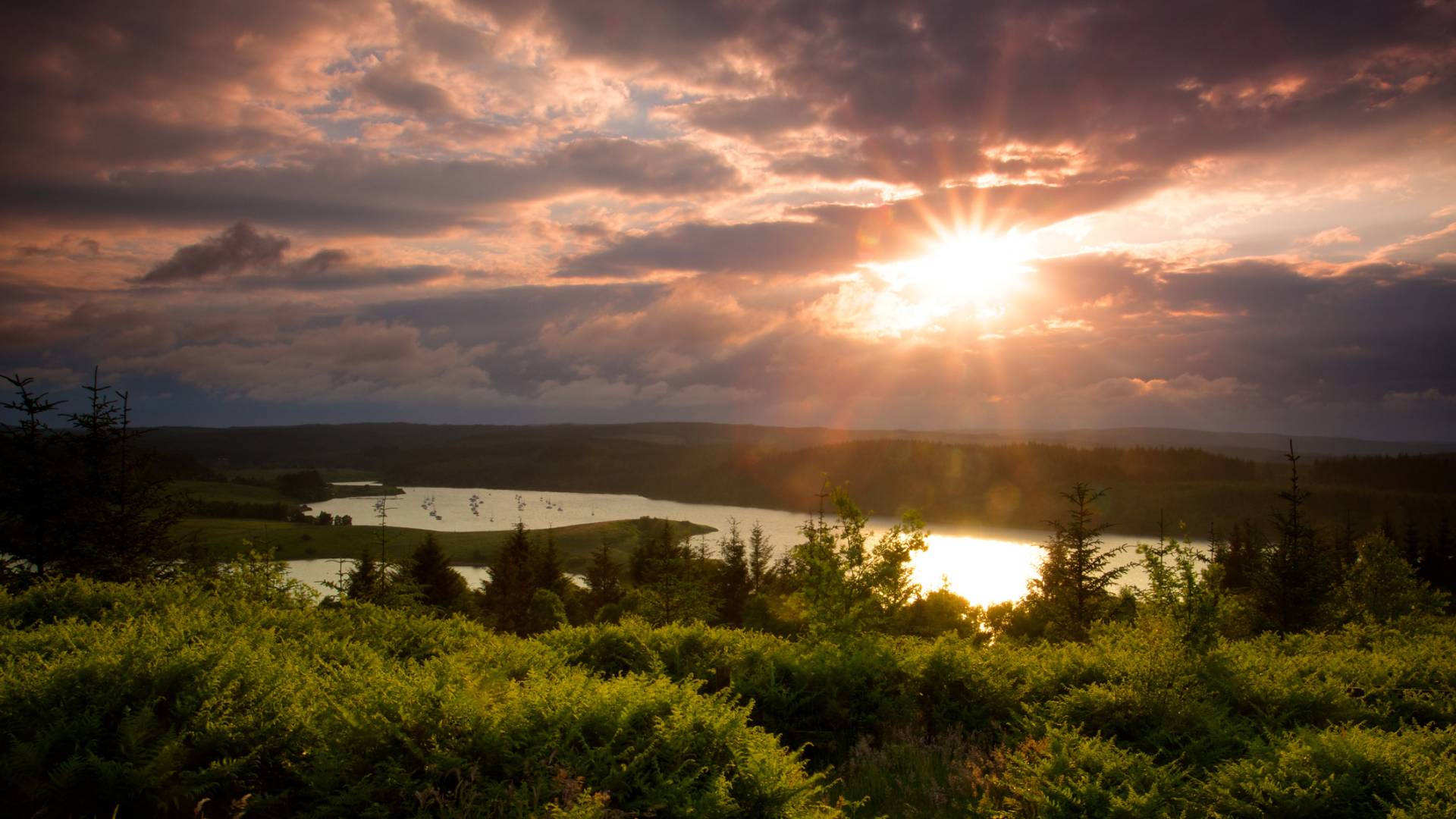 Sunset over a lush green landscape with a lake and trees; sunlight shines through dramatic clouds, casting warm light on the scene.