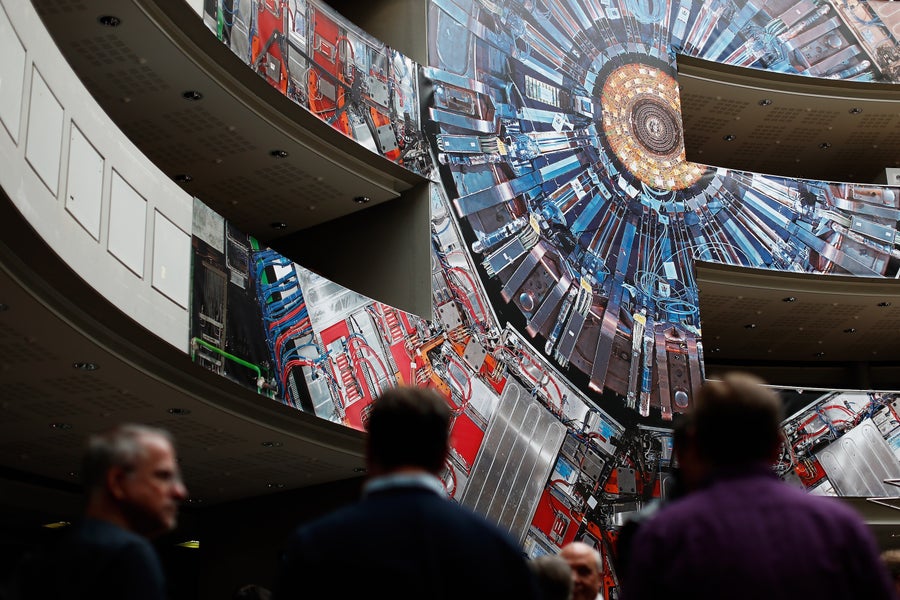 People stand in the foreground within a large building, with an enormous print of CERN's Compact Muon Solenoid experiment in the background.