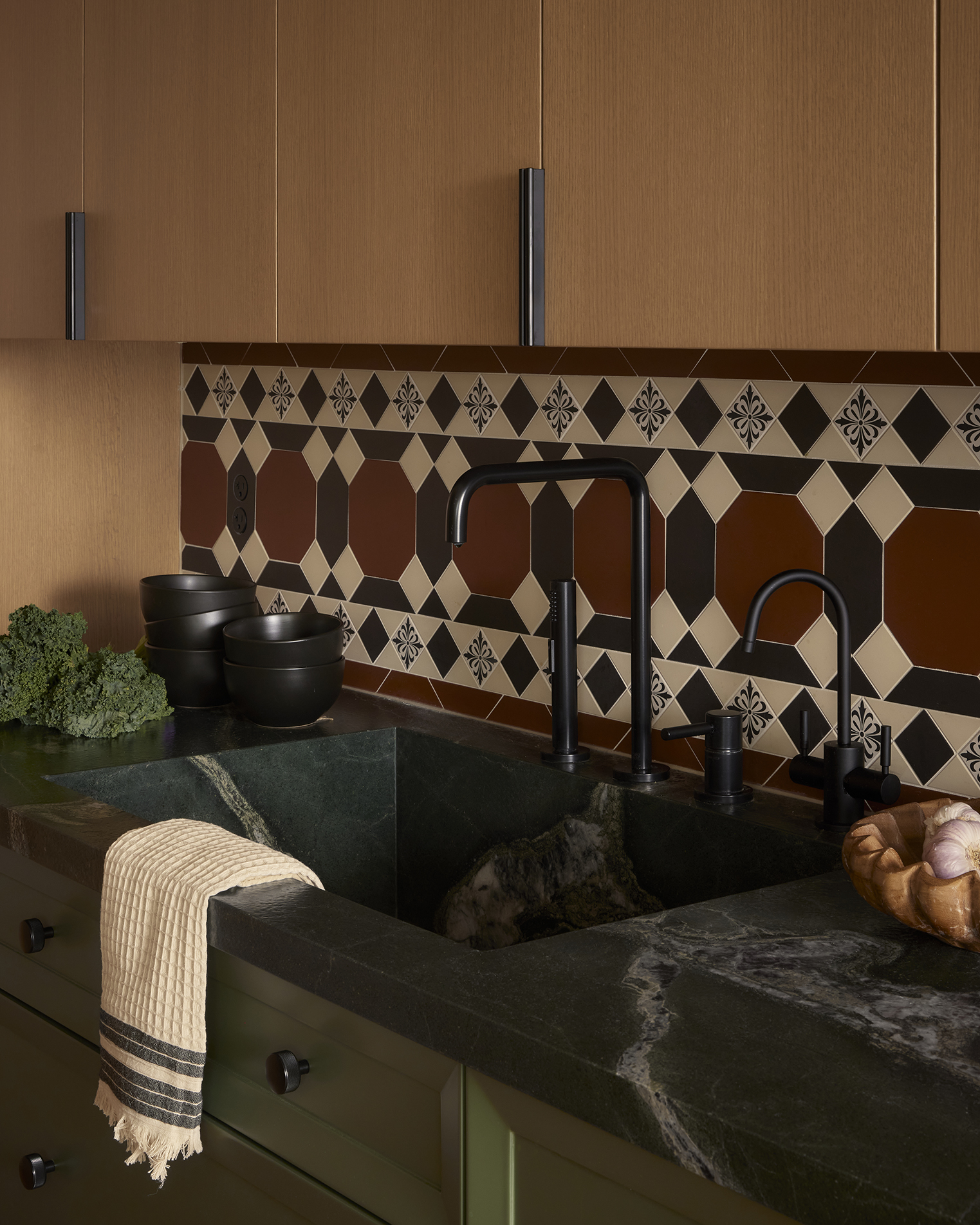 A nude and green wood kitchen with black marble countertops, tiled walls in black, beige, burned red, and blue, a series of dark earthenware bowls, fresh broccoli, and a tea towel.