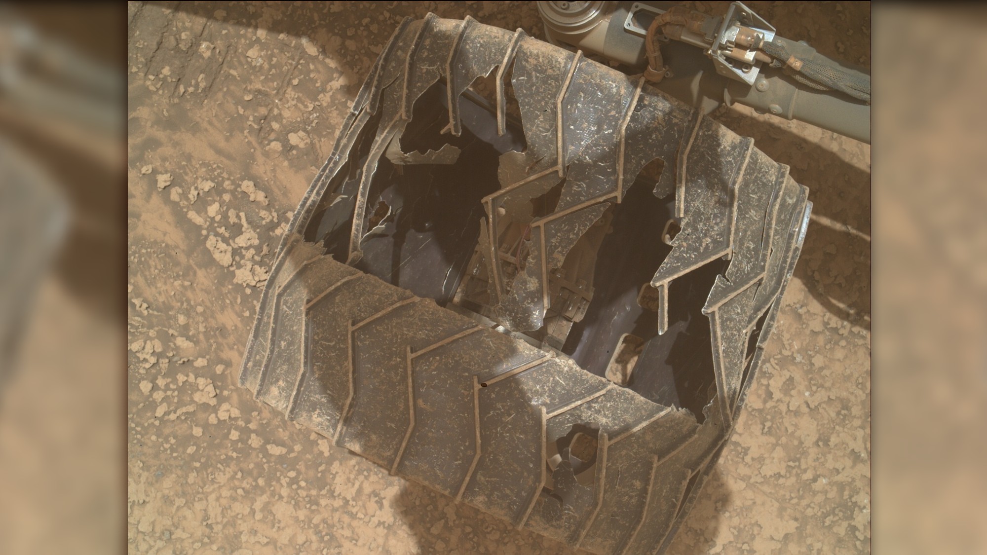 a close-up of a metal wheel with deep cracks and caved-in sections as it rests on a reddish-orange rocky surface