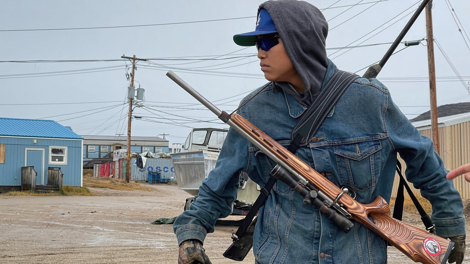 A bear guard in a hooded denim jacket and sunglasses stands watch with a rifle slung across his chest in the Arctic settlement of Kinngait.