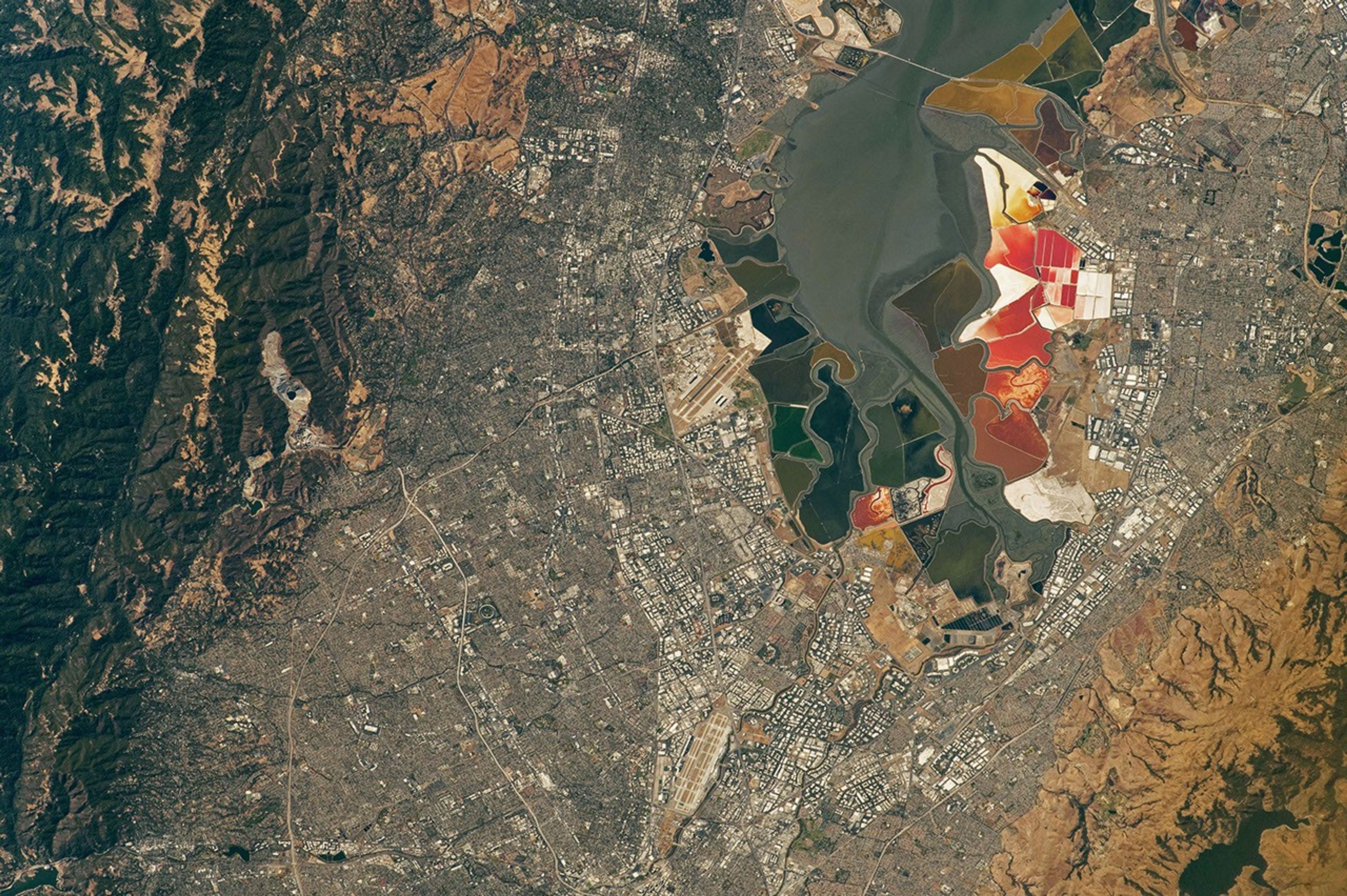 An aerial view of the San Francisco Bay Area Stadium, middle-left, is surrounded by urban infrastructure. Colorful ponds of the southern San Francisco Bay are visible toward the top.