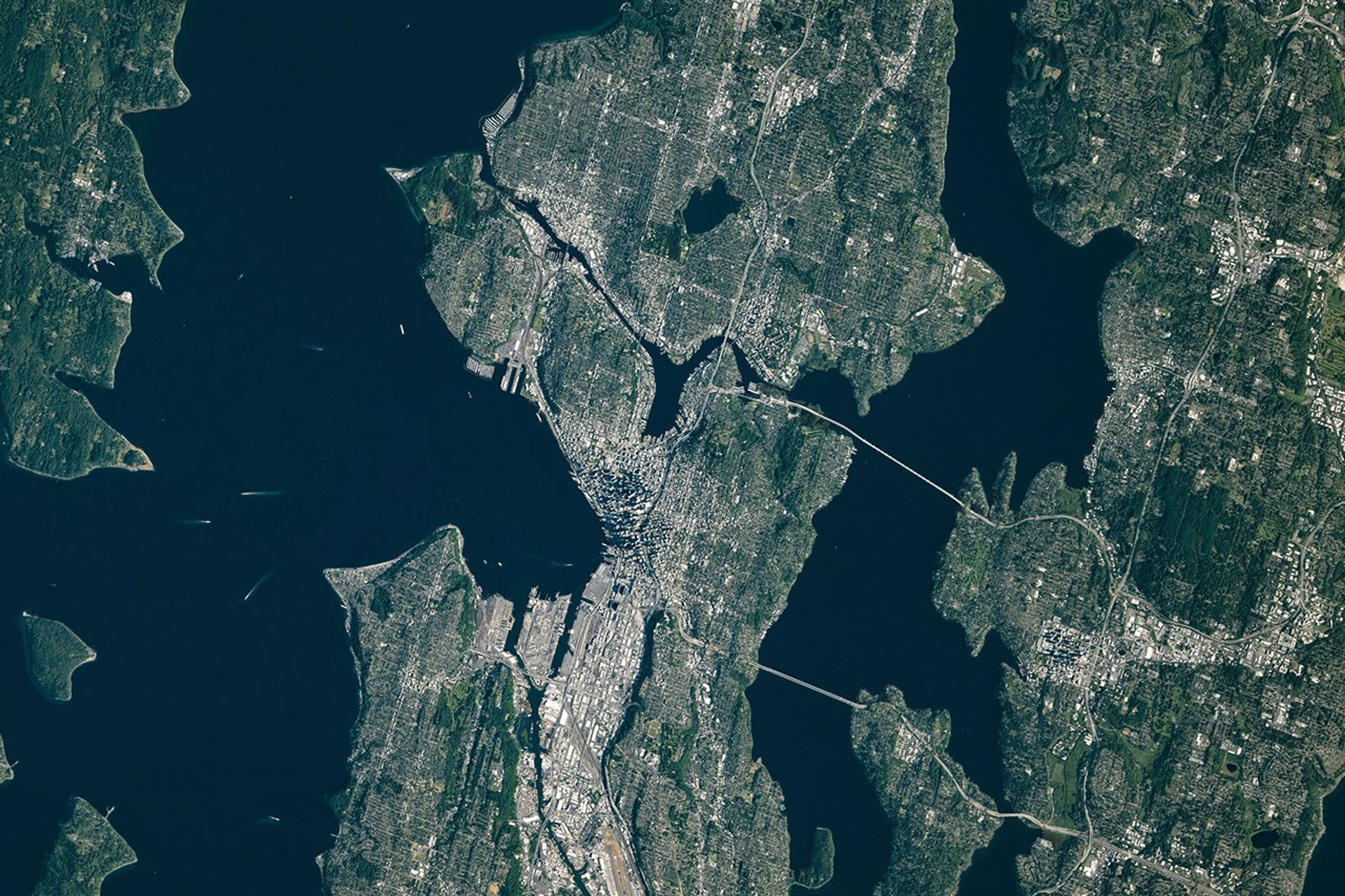 A top-down photo of the greater Seattle area shows a mix of light-colored developed areas and green parks and neighborhoods. Puget Sound and Lake Washington run along Seattle’s west and east sides, respectively.
