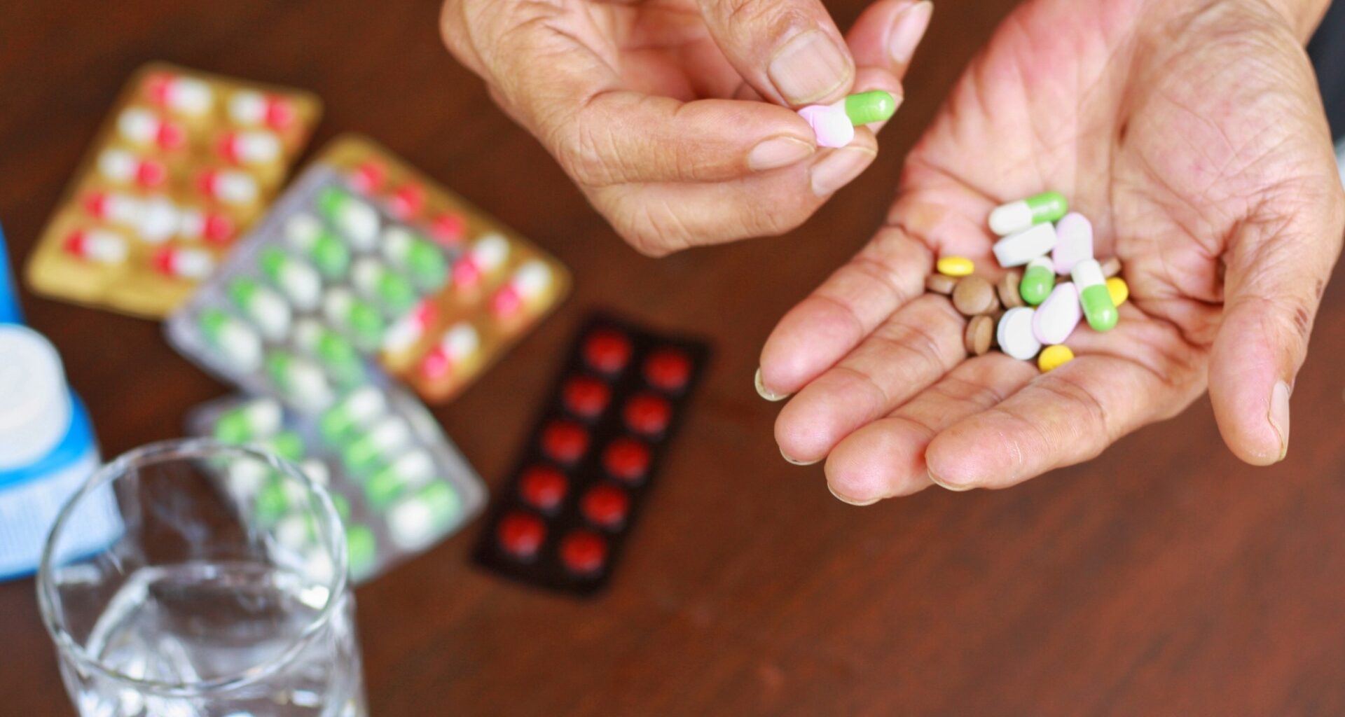 Elderly man hands holding many pills(tablet and capsule). Packets of mediation and glass of water