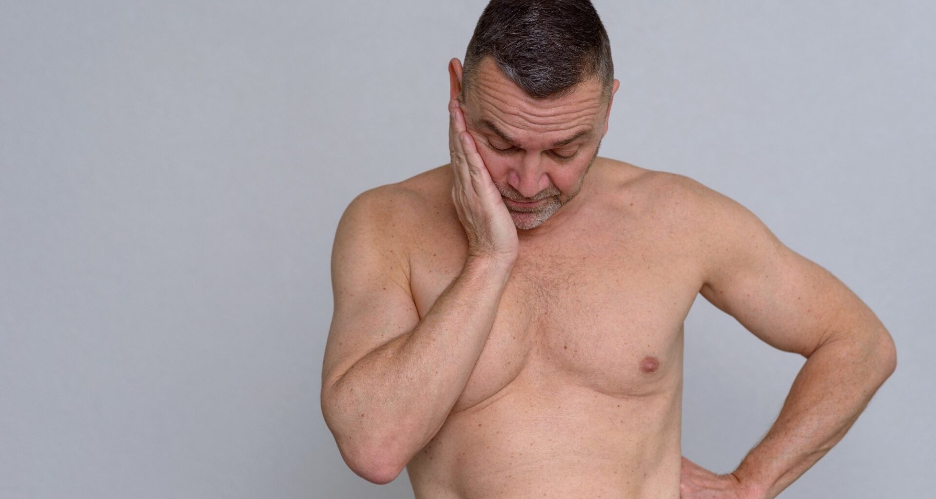 A studio portrait of a mature man naked from the waist up looking fed up and depressed.