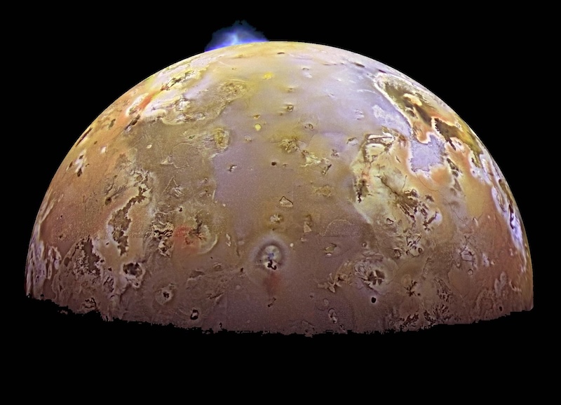 Planet-like body with mottled reddish-yellow surface and many spots. A small bluish plume shape is at the top.