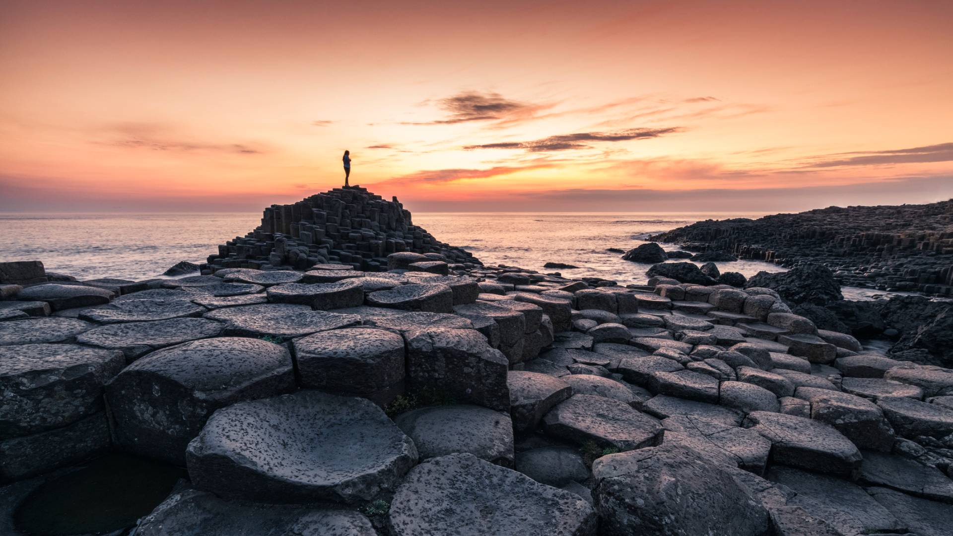 A person stands atop the hexagonal basalt columns of Giant&rsquo;s Causeway in Northern Ireland at sunset, with a colorful sky and calm sea in the background.