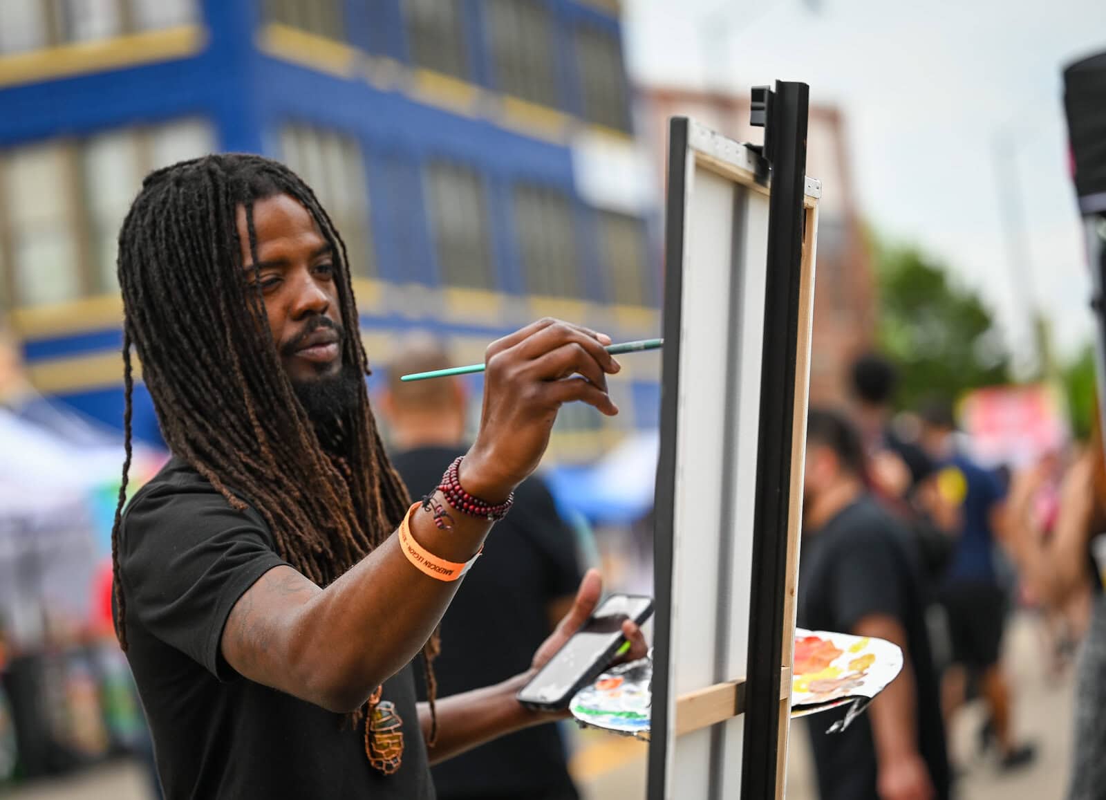 A man with long dreadlocks painting on an easel outdoors, holding a brush and palette with a crowd in the background.