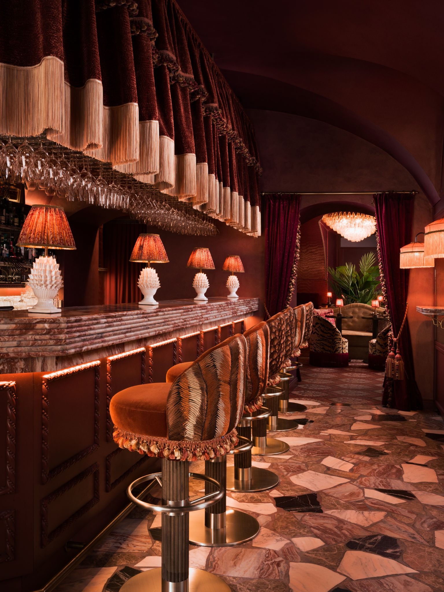The interiors of a plush cocktail bair with velvety red, golden, and green rooms decorated with glossy chandeliers, golden sconces, opulent drapery, and intricate patterns.