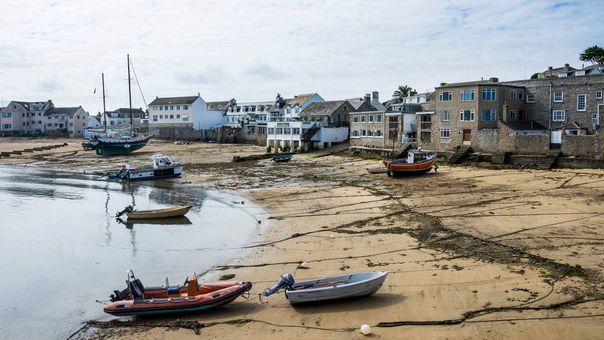 Small boats rest on the sandy shore of a coastal village at low tide, with waterfront buildings and a partly cloudy sky in the background.