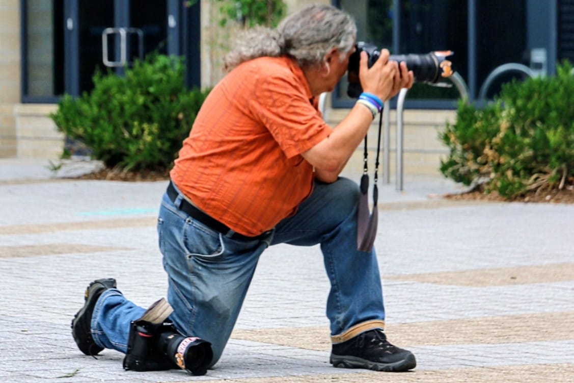 Person kneeling on one knee while using a DSLR camera to take a photo in a paved outdoor area.