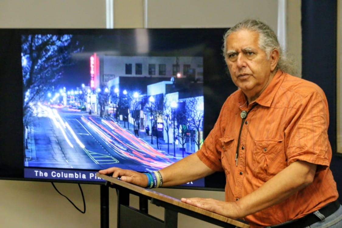 Man in an orange shirt stands at a lectern beside a large TV displaying a nighttime city street scene.