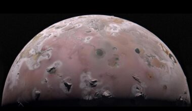 A view of half of the sphere of Jupiter’s volcanic moon Io against black space. The surface is colorful pinkish-tan and mottled with dark brownish features and sharp peaks surrounded by bright white diffuse deposits.