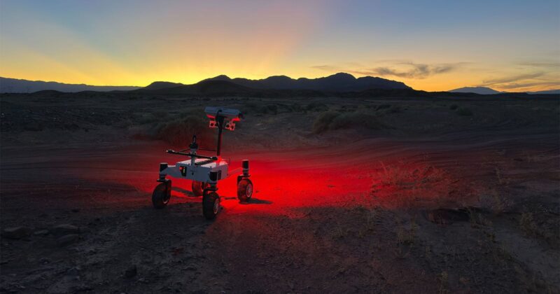 A small rover with lights glowing red underneath stands on rugged terrain at dusk, with mountains silhouetted against a colorful sunset sky in the background.