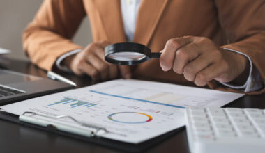 Person looking at documents with a magnifying glass at a desk with a laptop