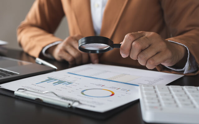 Person looking at documents with a magnifying glass at a desk with a laptop