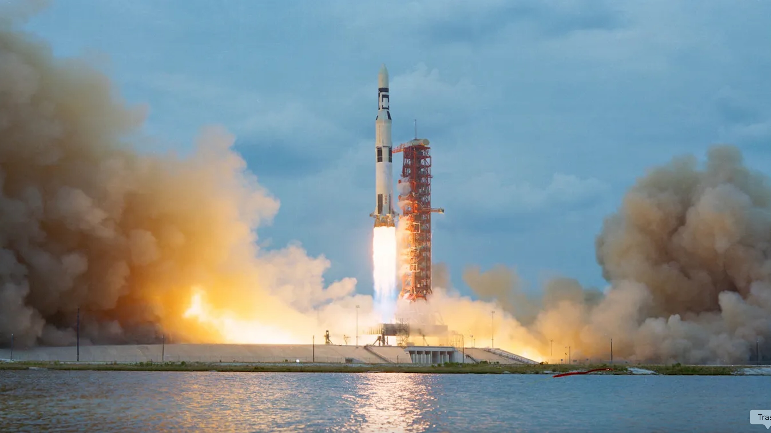 a big black and white rocket launches into a blue sky, with a lake in the foreground