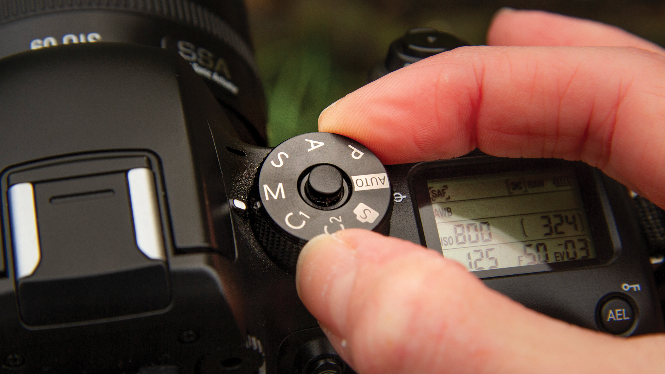 Hand adjusting mode dial on a DSLR camera set to various shooting modes with visible camera display settings