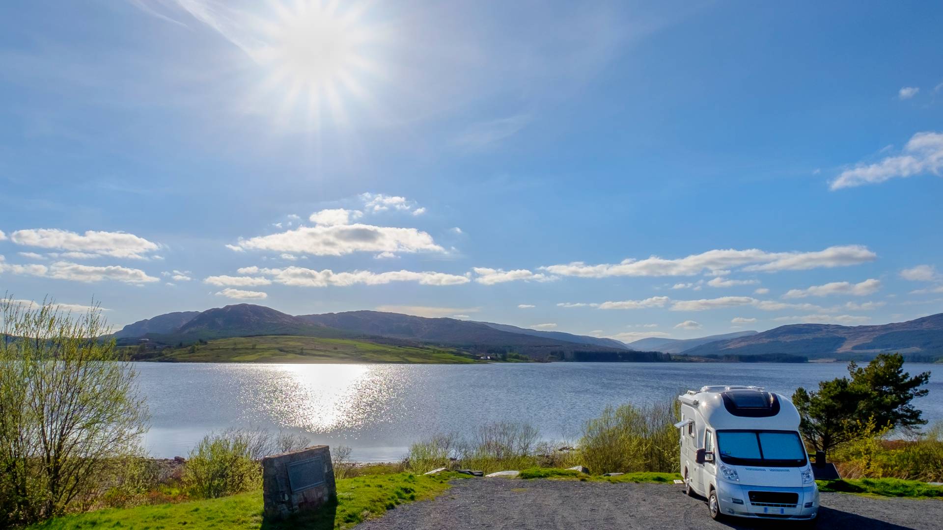A white camper van is parked by a calm lake with hills in the distance, under a bright, sunny sky with scattered clouds and the sun reflecting on the water.