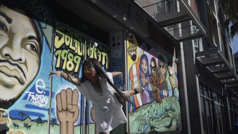 A person poses on one leg in front of a colorful mural featuring portraits, a raised fist, and text reading "SOLU WIDGETS 1989.