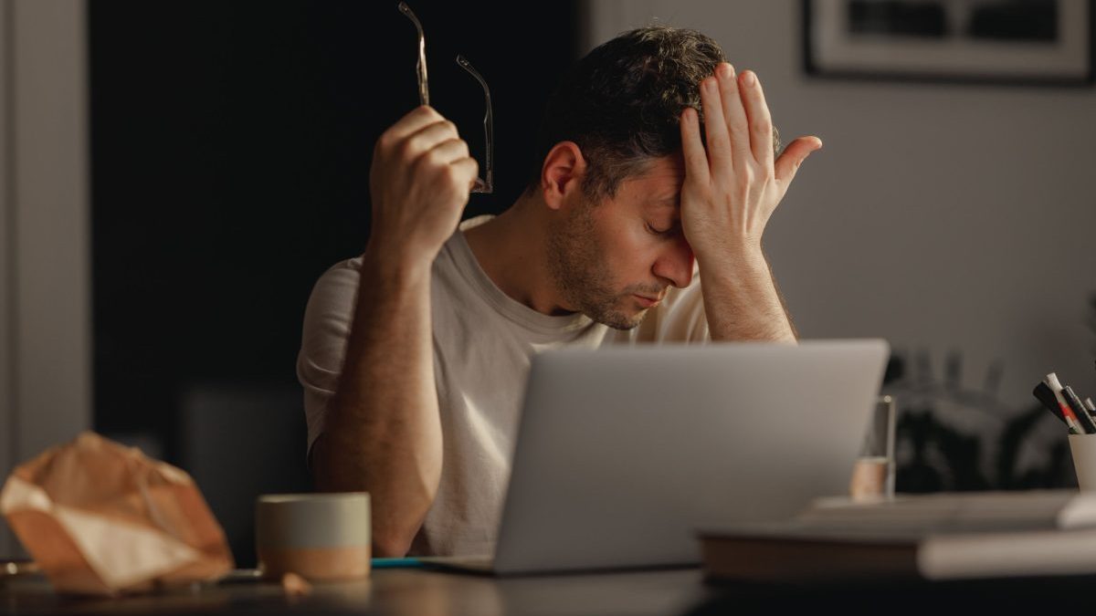 The image portrays a Caucasian man working on his laptop late at night at home, his expression and body language signaling stress and fatigue. The late hour adds to the atmosphere of intensity and pressure, highlighting the demanding nature of his work. This scene captures the challenging aspects of modern professional life, especially when working from home.