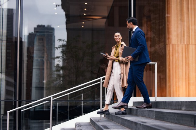 Two business professionals, a woman and a man, walk down exterior stairs beside a glass-fronted building. They converse and carry a tablet and folders, projecting a confident, city-work vibe.