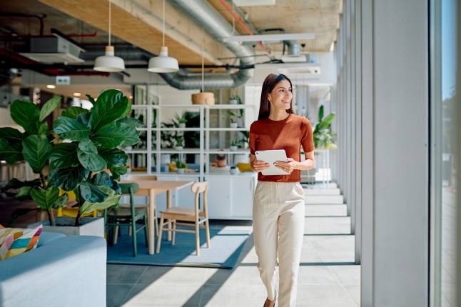 Businesswoman walking through a modern office, confidently holding a tablet and smiling while looking away, exuding professionalism and success