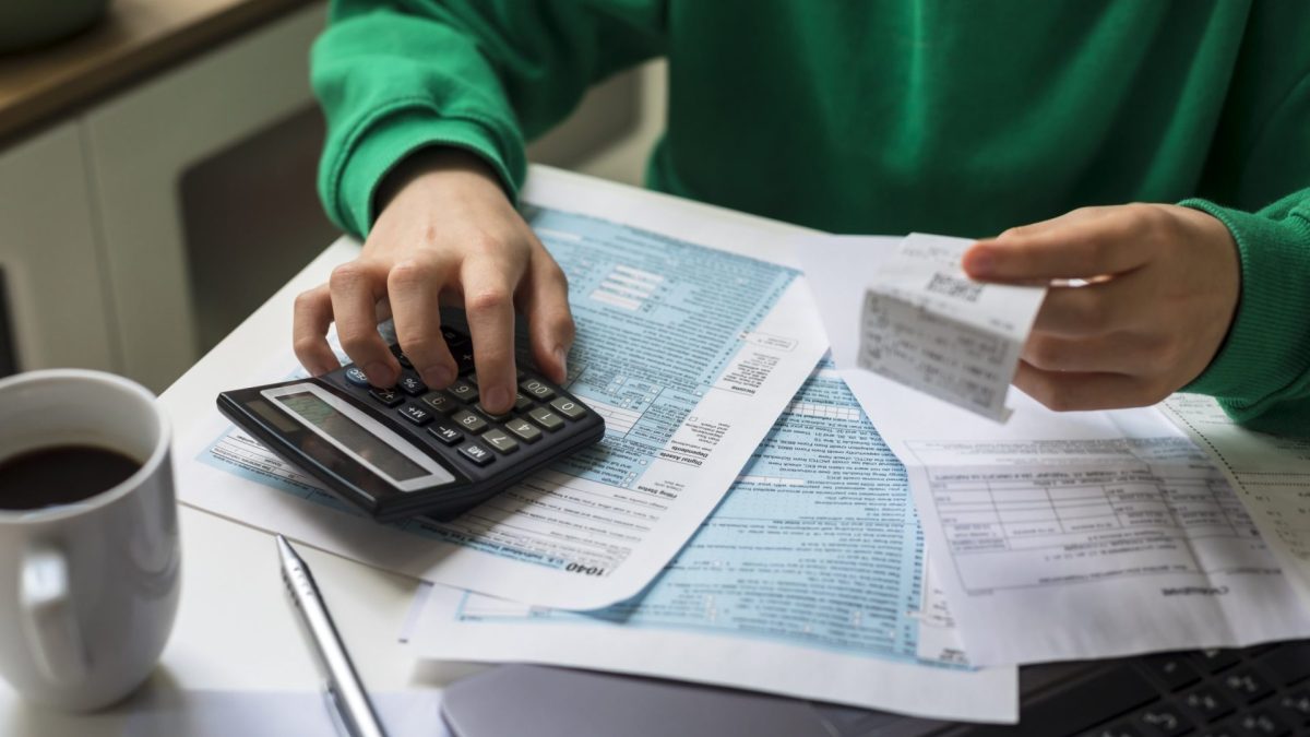 Person working at a home desk checking a receipt while entering amounts on financial paperwork with a calculator. Household finance organization, annual filing documents, tax preparation materials and general personal budgeting content.