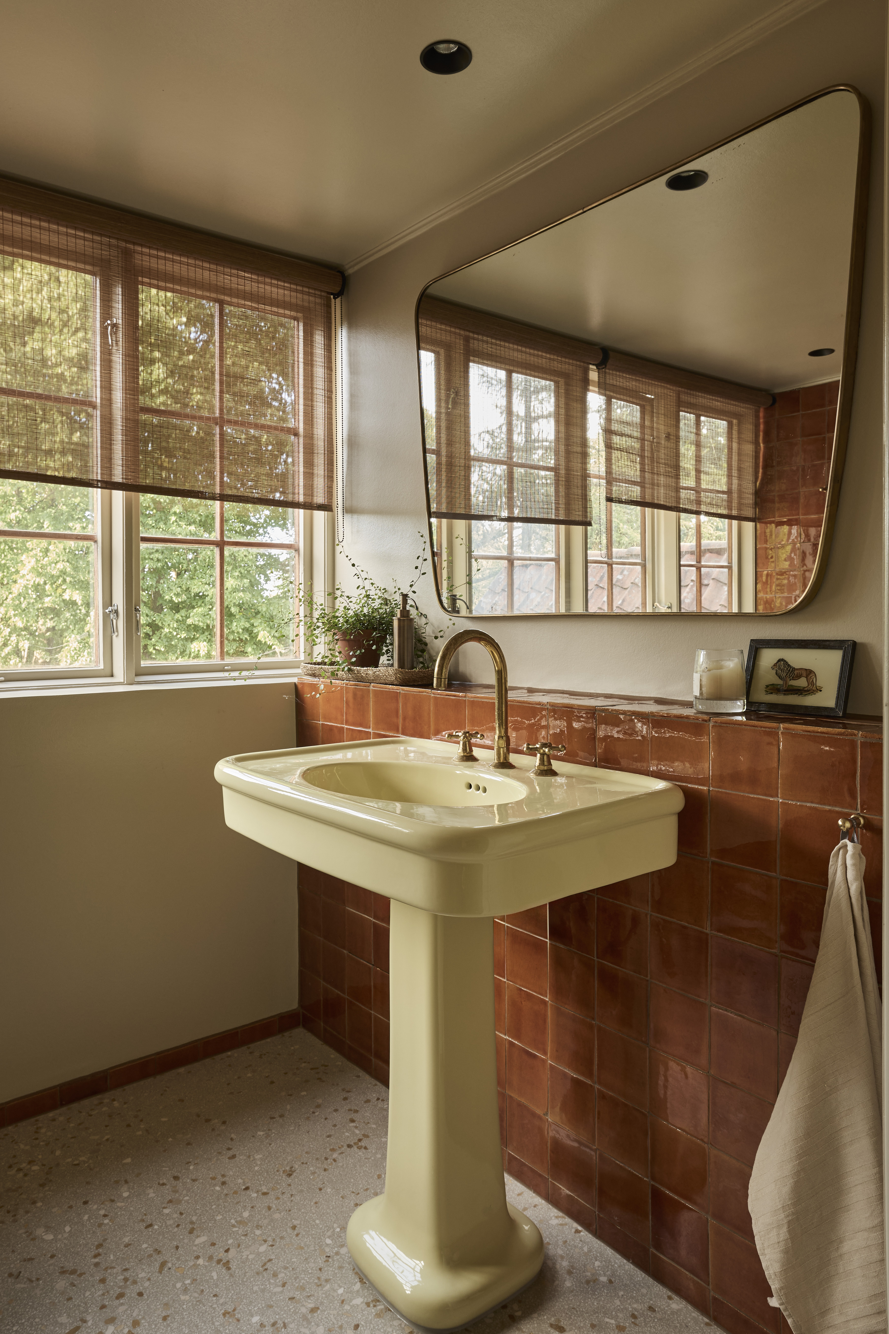 A butter yellow pedestal sink in a bathroom with terracotta tiles behind it creating a shelf for decor underneath a large mirror