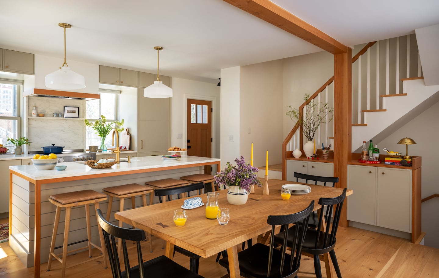 A kitchen with a rectangular table in the foreground with six chairs. Behind that, there's an island with four stools. To the right, there are stairs leading up.