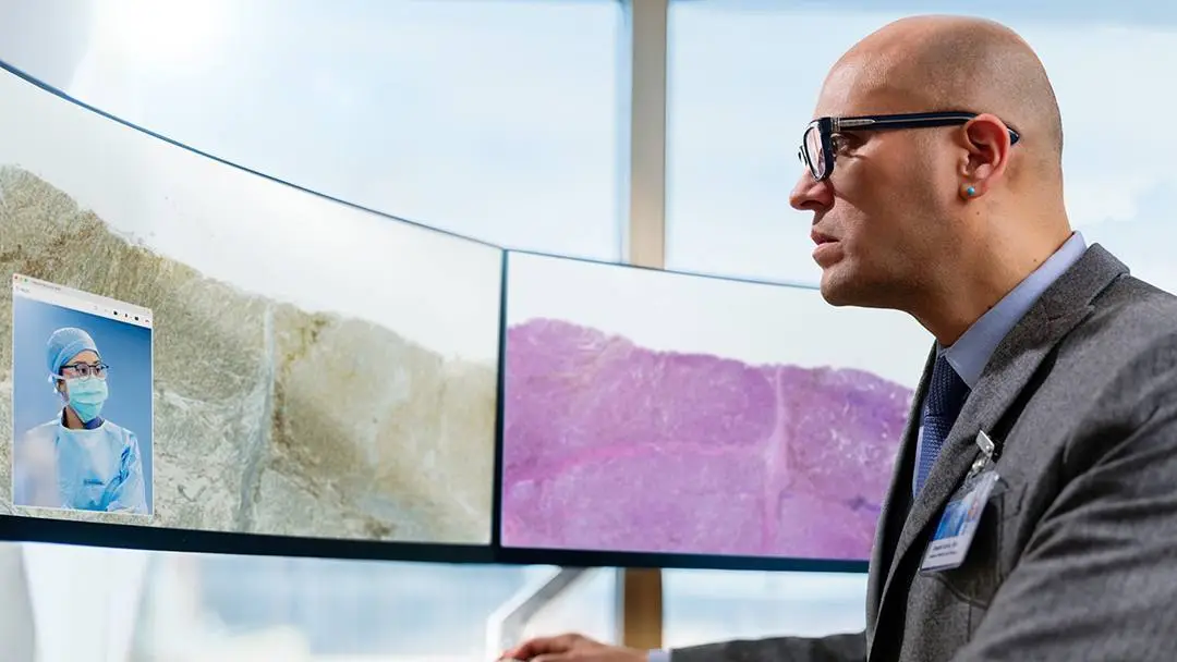 A healthcare professional reviews digital pathology images on three large monitors while consulting remotely with a colleague via video call, illustrating digital pathology and telemedicine.