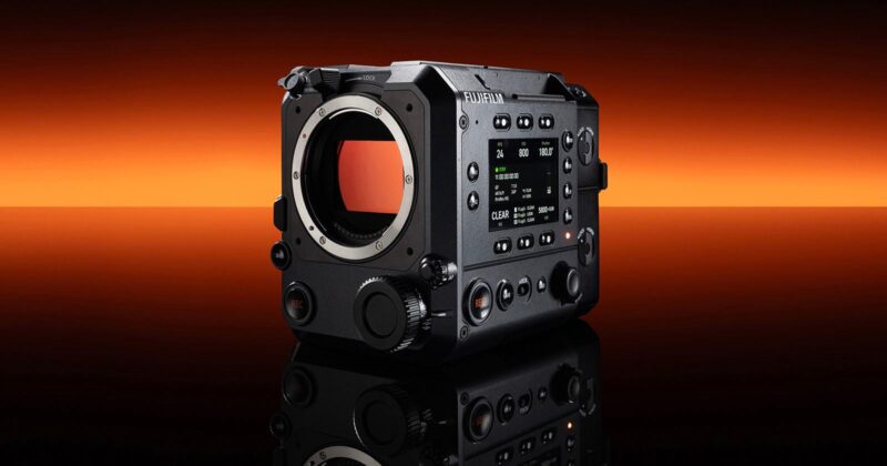 A black Fujifilm digital cinema camera body is displayed on a reflective surface with an orange to black gradient background. The camera features multiple dials, buttons, and a digital screen on its side.