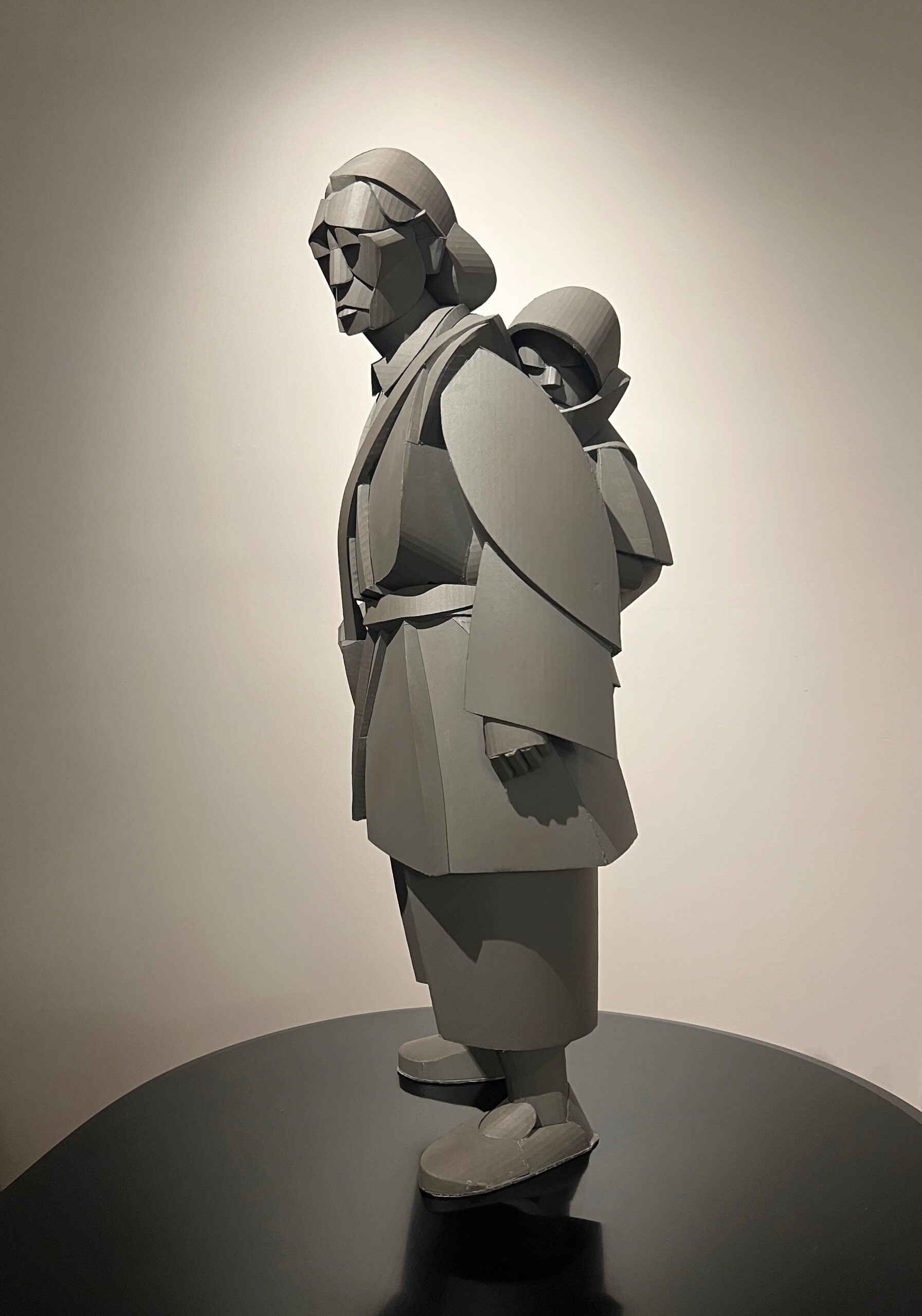 Gray geometric sculpture of a person in traditional clothing carrying a child on their back, displayed on a black pedestal against a plain wall.