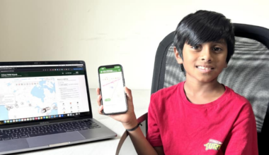 A young boy sits in front of a laptop and is holding a phone. On the computer is a map of Canada with certain places indicated.