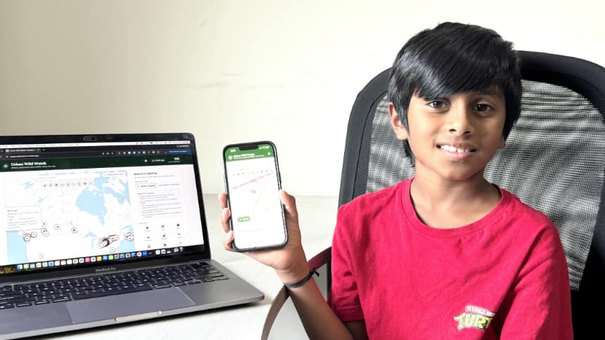 A young boy sits in front of a laptop and is holding a phone. On the computer is a map of Canada with certain places indicated.
