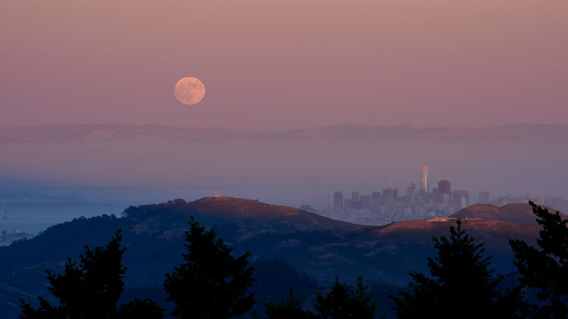 A full moon rises in a peach colored sky over the hills near a city, whose skyscrapers can be seen rising to the right of the photo.