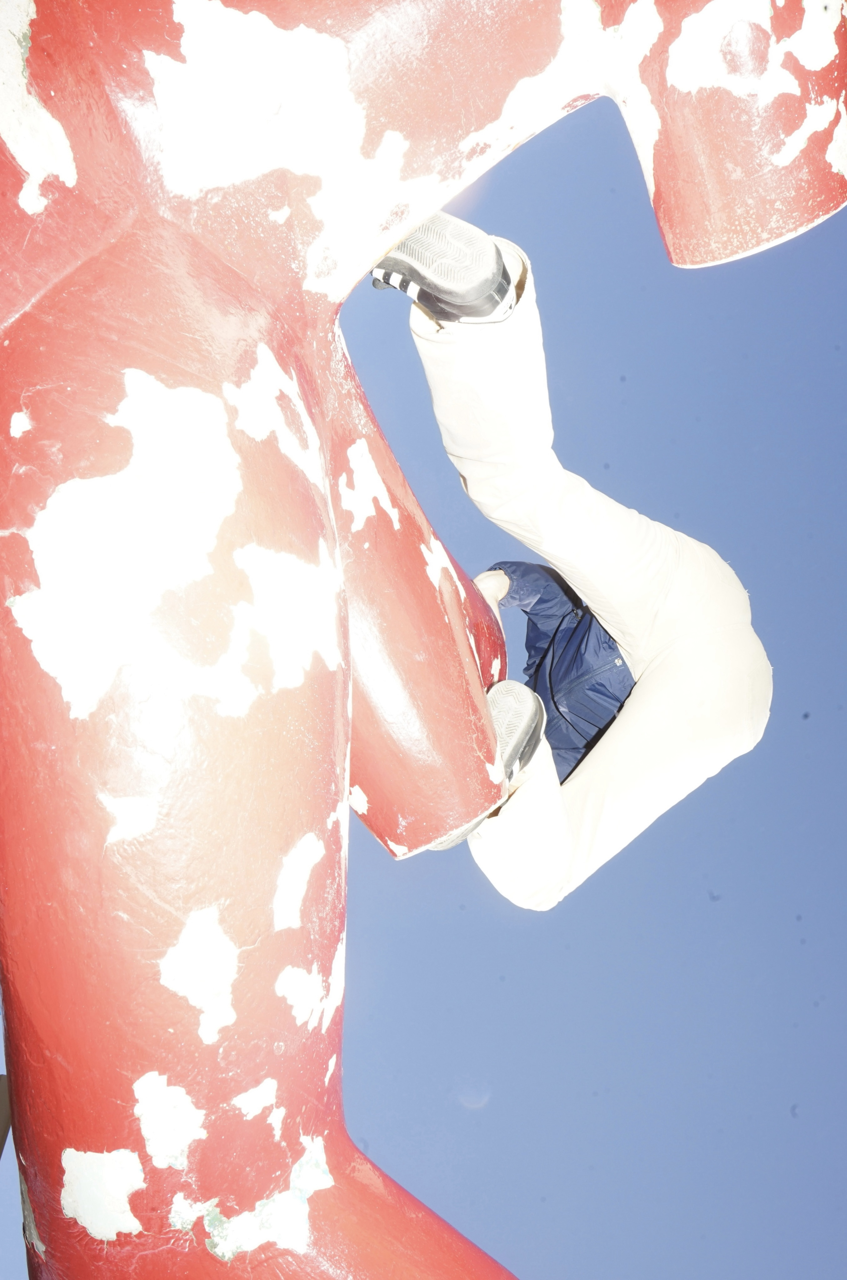 An individual wearing a blue jacket and white pants climbs horizontally across a large, weathered red sculpture with peeling paint against a clear blue sky.