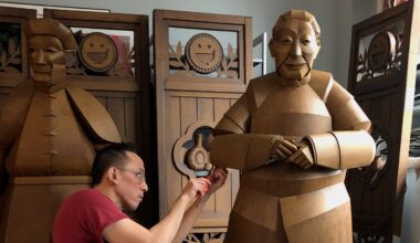 A person in a red shirt works on a large, brown, human-like sculpture in a room with wooden folding screens and another similar sculpture in the background.