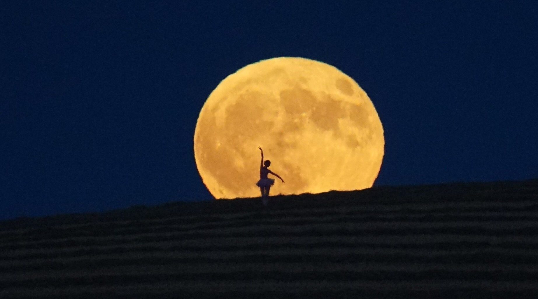 The silhouette of a ballerina is shown dancing in front of the disk of a full moon as it rises over a hillside in a dark evening sky.