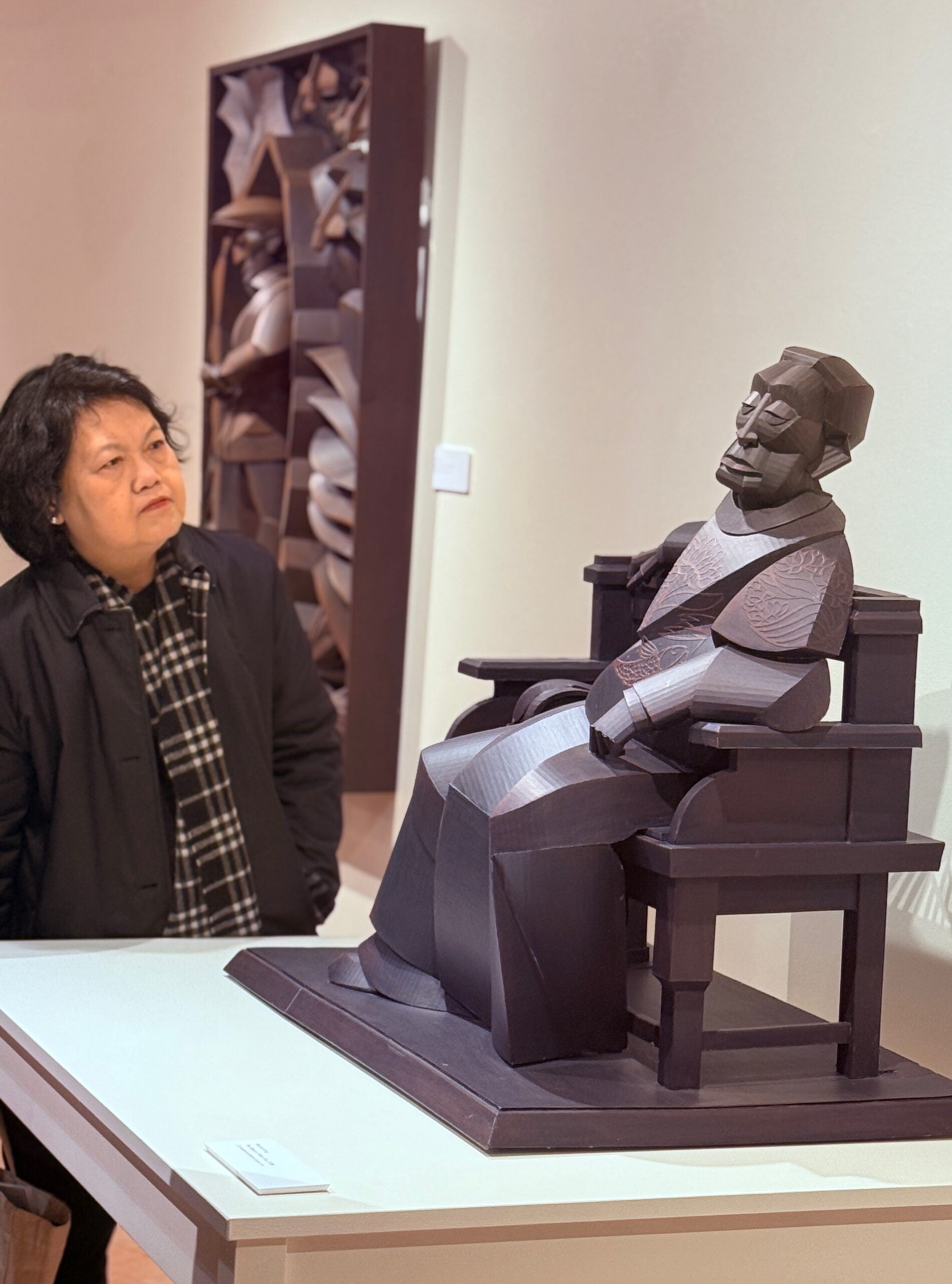 A woman looks at a wooden sculpture of a seated figure displayed on a white pedestal in an art gallery.