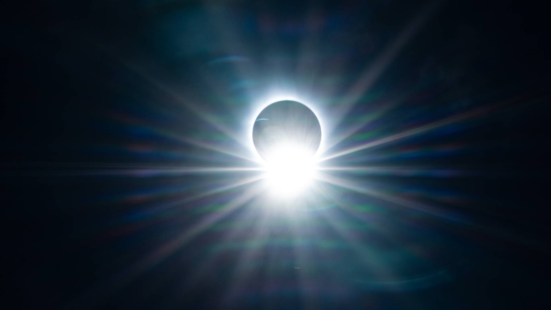 A total solar eclipse. The black disc of the moon has almost completely covered the sun. The moon is surrounded by a glowing, wispy white ring of light against the dark night sky.