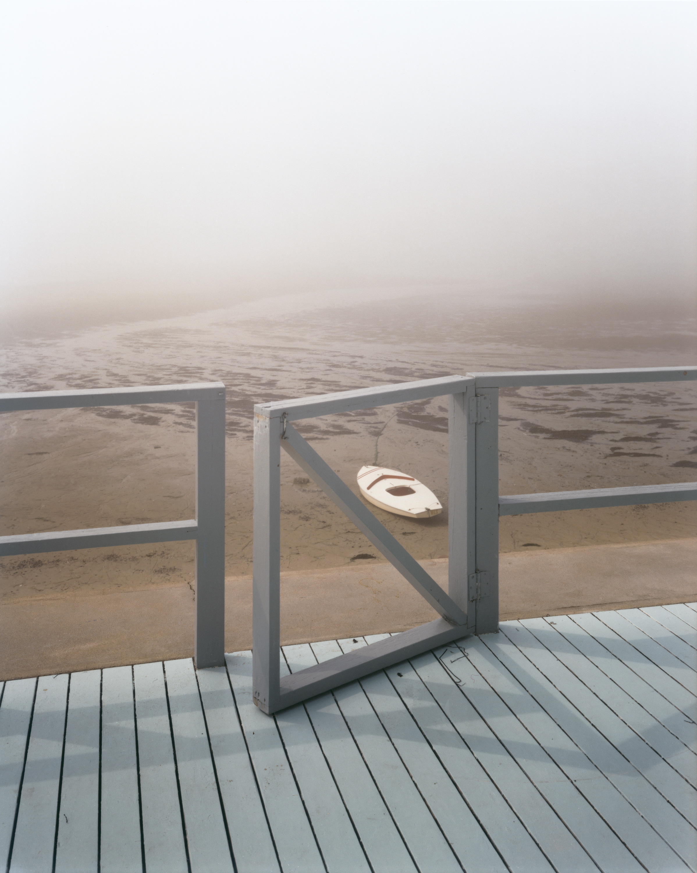 A weathered wooden gate opens onto a fog-shrouded beach, where a small white sailing dinghy sits stranded on the wet sand.