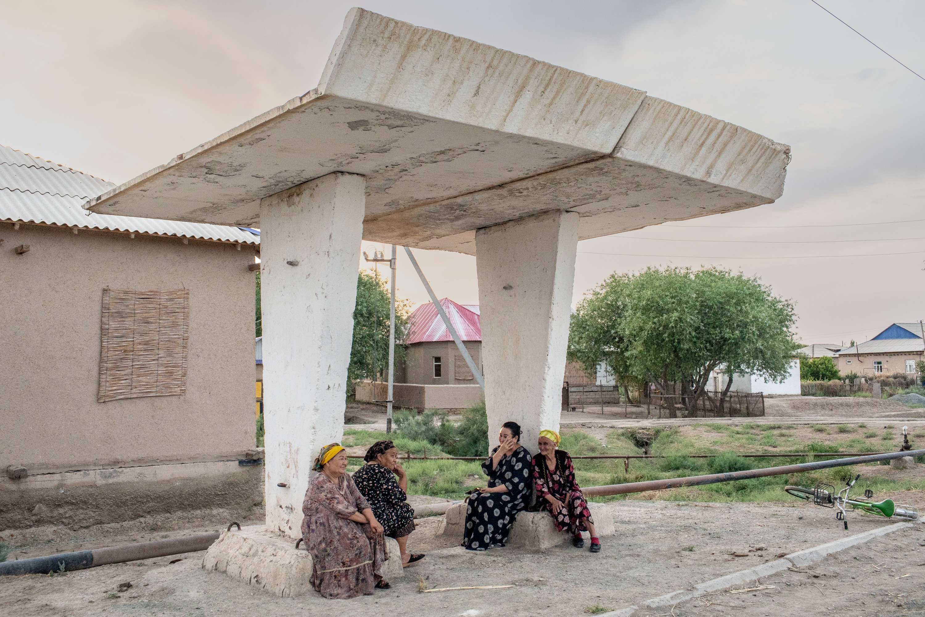 Four women sit and converse in the shade under a large concrete, canopy-like structure in an open, rural landscape with a bicycle on the ground nearby.