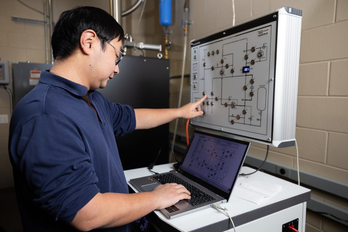 Han Zhao pointing at a control panel while using a laptop, showing hands-on setup and data review for superconducting and nanomechanical experiments.